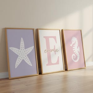 May include: Three framed prints with a coastal theme. One features a white starfish on a lavender background. Another displays a pink letter "E" with the name "emma" in cursive. The third print shows a white seahorse on a pink background.