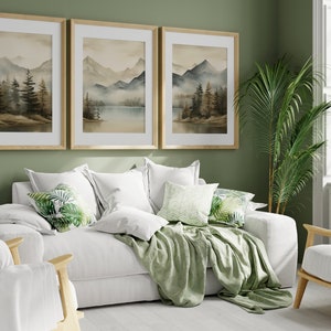 May include: A white couch with green throw pillows and a green throw blanket in a living room with a green wall, three framed watercolor paintings of mountains and a lake, a potted plant, and a white chair with a green throw pillow.