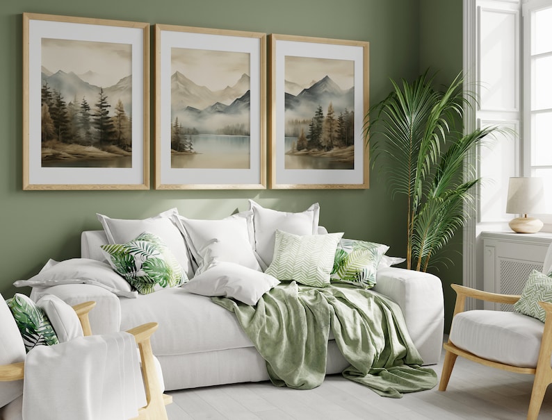 May include: A white couch with green throw pillows and a green throw blanket in a living room with a green wall, three framed watercolor paintings of mountains and a lake, a potted plant, and a white chair with a green throw pillow.