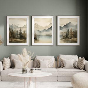 May include: Three framed watercolor paintings of a misty mountain landscape with a lake in the middle. The paintings are hung on a sage green wall above a light beige sofa with pillows.