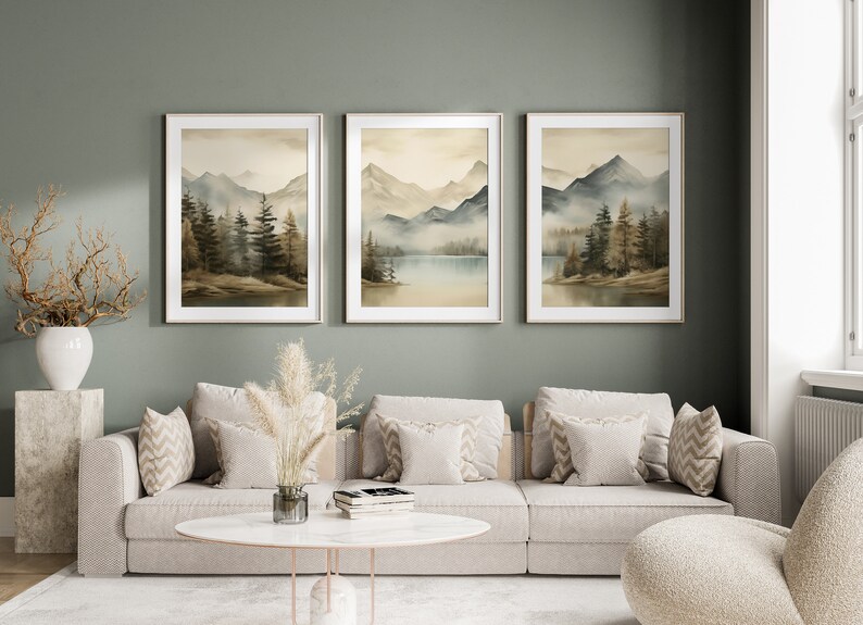 May include: Three framed watercolor paintings of a misty mountain landscape with a lake in the middle. The paintings are hung on a sage green wall above a light beige sofa with pillows.