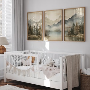 May include: A white crib with a beige blanket and pillows, decorated with a garland of fabric flags. Three framed landscape paintings of mountains and a lake hang on the wall behind the crib.