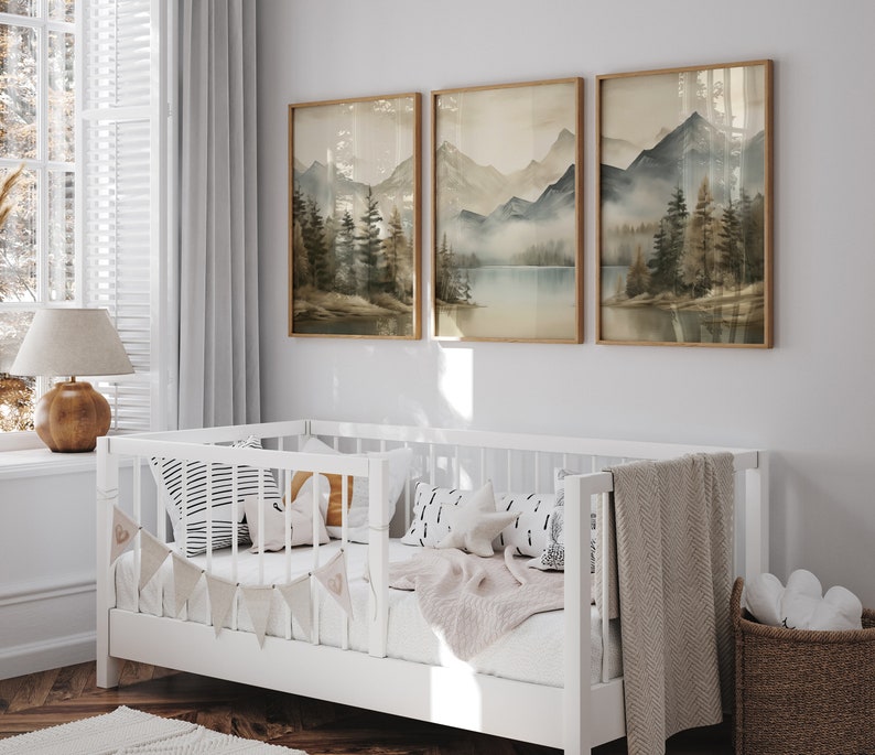 May include: A white crib with a beige blanket and pillows, decorated with a garland of fabric flags. Three framed landscape paintings of mountains and a lake hang on the wall behind the crib.