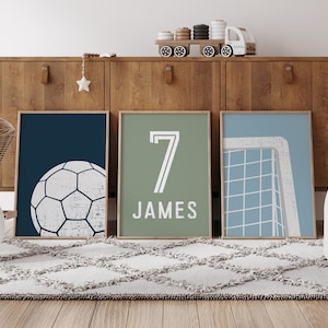Könnte beinhalten: Drei gerahmte Drucke mit einem Sportthema. Der erste Druck hat einen dunkelblauen Hintergrund mit einem weißen Fußball. Der zweite Druck hat einen grünen Hintergrund mit der Nummer 7 und dem Namen James. Der dritte Druck hat einen hellblauen Hintergrund mit einem weißen Fußballtor.