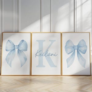 Op de afbeelding: Drie ingelijste prints met een lichtblauw strikdesign en een gepersonaliseerde print met de naam "Keilani" en de letter "K". De prints zitten in gouden frames en worden op een houten vloer getoond.