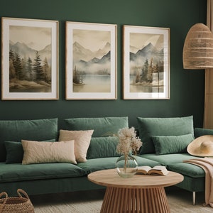 May include: Three framed watercolor paintings of a misty mountain landscape with a lake in the foreground. The paintings are hung on a dark green wall above a green sectional sofa with pillows and a throw blanket.