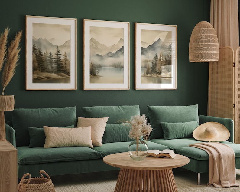 May include: Three framed watercolor paintings of a misty mountain landscape with a lake in the foreground. The paintings are hung on a dark green wall above a green sectional sofa with pillows and a throw blanket.