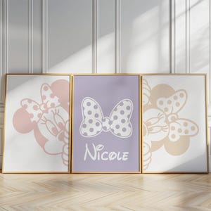 May include: Three framed art prints with gold frames. The prints feature Minnie Mouse designs in pink, beige, and purple. The center print has a bow design with the name "Nicole" below it. The prints are displayed on a wooden floor.