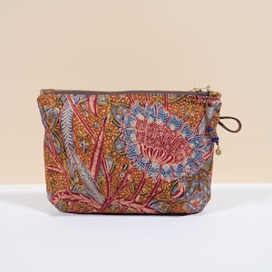 Könnte beinhalten: Eine rechteckige Kosmetiktasche mit einem Blumenmuster in Braun-, Rot- und Blautönen. Die Tasche hat einen Reißverschluss und eine kleine Lederschlaufe mit blauen Perlen und einem Anhänger. Die Tasche steht auf einer weißen Oberfläche.