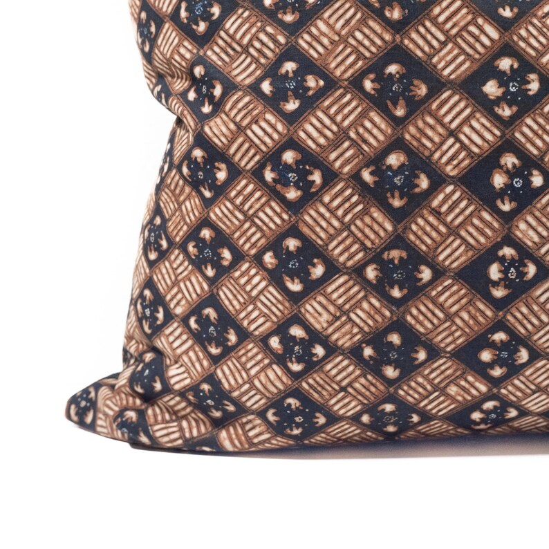 May include: Close-up of a decorative pillow with a repeating geometric pattern. The pattern features brown and navy blue diamond shapes with floral-like accents. The pillow has a soft, textured appearance and is set against a white background.