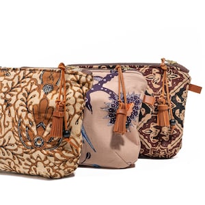 May include: Three fabric pouches with zip closures and leather tassel accents. The pouches feature unique patterns in shades of brown, beige, and purple. The pouches are designed for carrying small items.