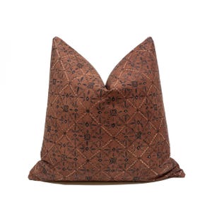 Peut inclure: Un coussin carré de couleur brun terre cuite sourd présentant un motif en grille de losanges répétitifs. Le design comprend des motifs floraux gris anthracite centrés dans chaque losange, accentués par de petits points blancs sur les lignes.