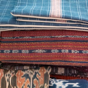 May include: A stack of colorful woven fabric with different patterns and textures. The top layer is a light blue fabric with a white and pink stripe. The second layer is a dark blue fabric with a white stripe. The third layer is a red fabric with a blue and yellow stripe. The bottom layer is a dark blue fabric with a white stripe.