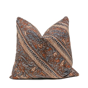 Puede incluir: Un cojín decorativo con un estampado batik marrón, azul y crema. El cojín presenta un diseño diagonal con elementos florales y geométricos. La tela tiene una apariencia texturizada, agregando interés visual al artículo de decoración del hogar.
