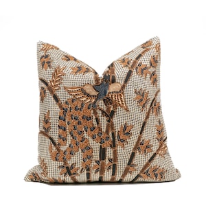 Op de afbeelding: Vierkant decoratief kussen met een batikontwerp. Het heeft een crèmekleurige achtergrond met een rasterpatroon. Het ontwerp omvat bruine bamboestengels, bladeren en een vogel met blauwe en bruine accenten. Het kussen is vierkant van vorm.