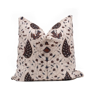 Puede incluir: Cojín decorativo color crema con un detallado estampado batik. El diseño presenta motivos florales y de aves en marrón, azul marino y crema. El cojín es de forma cuadrada y tiene un aspecto suave y texturizado.