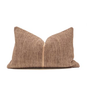 Puede incluir: Un cojín decorativo rectangular con un patrón de rayas marrones y beige. El cojín tiene un diseño de rayas verticales con una raya beige más clara en el centro. El cojín está hecho de tela texturizada.
