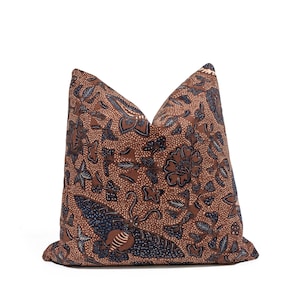 May include: A square throw pillow featuring a brown fabric base with a dark blue and white batik-style pattern. The design includes intricate floral motifs, leafy vines, and a prominent moth or butterfly graphic centered on the lower portion.