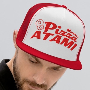 以下が含まれることがあります： 赤と白のトラッカーハット。赤色の「Mama's Pizza ATAMI」の文字入り。つばは赤色で、前面パネルは白色にグラフィック入り。黒いジャケットを着た人がかぶっています。