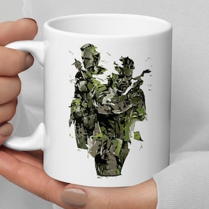 May include: White ceramic mug with a graphic of three characters from a video game. The characters are depicted in a camouflage pattern and are holding weapons.