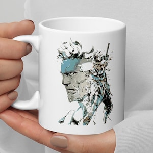 May include: White ceramic mug with a graphic of a man's face with a bandana and a camouflage pattern. The man is looking to the right.