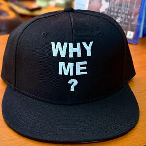 May include: Black baseball cap with a flat brim, featuring the white text "WHY ME?" embroidered on the front. The cap rests on a wooden surface. A video game case is visible in the background.