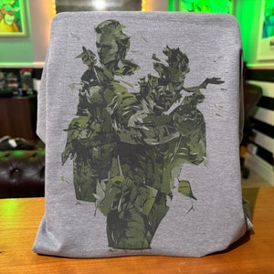 May include: A grey t-shirt featuring a camouflage print of three figures. The figures are in a fighting stance, with their faces obscured by shadows.