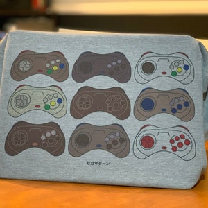May include: A gray t-shirt with a design of nine different colored video game controllers. The controllers are all in a row and are all facing the same direction. The text "セガサターン" is printed below the controllers.