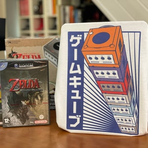 May include: Three video game cases for the Nintendo GameCube, featuring the game "The Legend of Zelda: Twilight Princess". The cases are on a wooden table next to a white tote bag with a graphic of a stack of GameCube controllers and the Japanese text "ゲームキューブ".