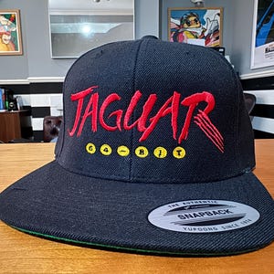 May include: Black and red baseball cap with the word "JAGUAR" embroidered in red on the front. The cap also has the words "64-BIT" embroidered in yellow below the word "JAGUAR".