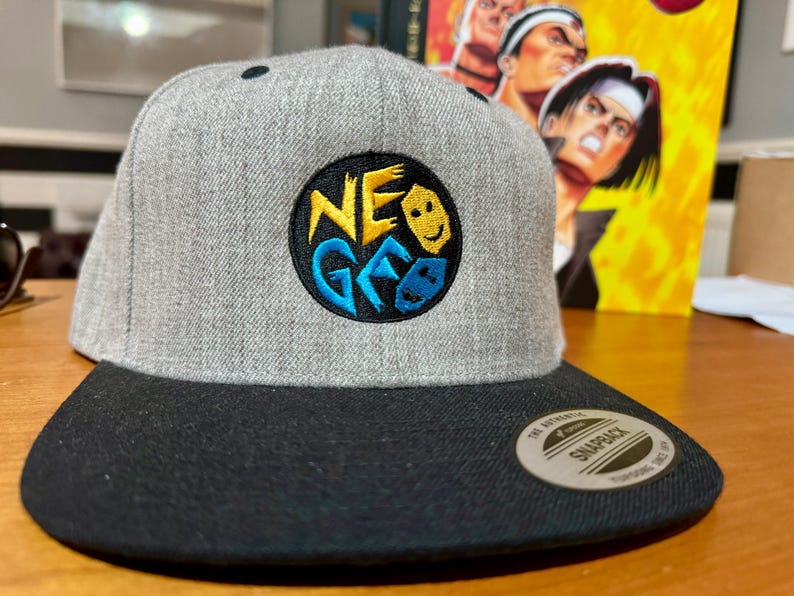 May include: A gray and black baseball cap with a black and yellow embroidered logo that says "NEO GEO" with a blue and yellow smiley face design.