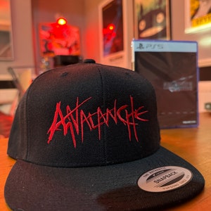Könnte beinhalten: Schwarze und rote Baseballkappe mit dem Wort "Avalanche" in rot auf der Vorderseite gestickt. Die Kappe hat einen schwarzen verstellbaren Riemen mit einem weißen "The Authentic Yupoong Snapback"-Etikett.