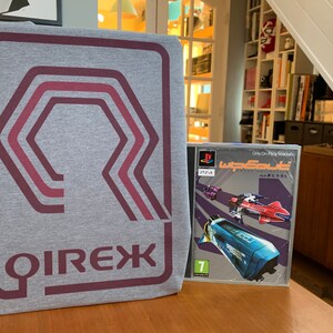 Może przedstawiać: Szary T-shirt z bordowym logo przedstawiającym stylizowany wzór geometryczny i napis "QIREX" nadrukowany od tyłu. Obok T-shirtu znajduje się pudełko gry PlayStation "Wipeout XL" z srebrnym tłem i kolorowymi statkami kosmicznymi.