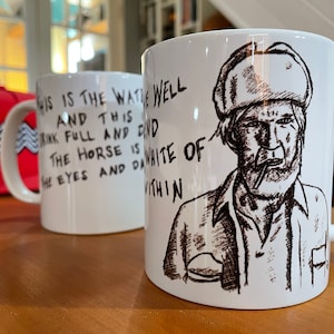 May include: White ceramic mug with a black and white illustration of a man with a beard and a hat. The text on the mug reads "THIS IS THE WATIE WELL AND THIS RINK FULL AND DID THE HORSE IS THE EYES AND DI WHITE OF VITHIN".