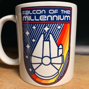 Puede incluir: Taza de cerámica blanca con un gráfico azul, naranja y amarillo del  Millennium Falcon. El texto "FALCON OF THE MILLENNIUM" está impreso en azul en la taza.