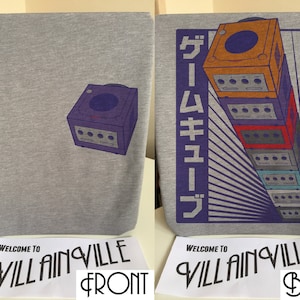 Może przedstawiać: Szary T-shirt z nadrukowaną na przodzie fioletową konsolą GameCube i stosem konsol GameCube z japońskim tekstem "ゲームキューブ" nadrukowanym na plecach. Tekst "WELCOME TO VILLAINVILLE" jest nadrukowany na białej metce pod koszulą.