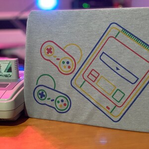 Op de afbeelding: Een grijs T-shirt met een retro-illustratie van een Super Nintendo Entertainment System-console en twee controllers. De console is omlijnd in geel, groen en rood, terwijl de controllers zijn omlijnd in groen en blauw.