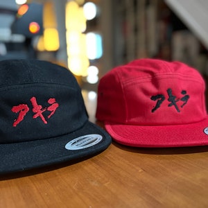Op de afbeelding: Twee zwarte en rode baseballpetten met het Japanse woord "Akira" in rood geborduurd op de voorkant van elke pet.