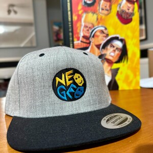May include: A gray and black baseball cap with a black and yellow embroidered logo that says "NEO GEO" with a smiley face and a video game controller graphic. The cap is a snap back style.