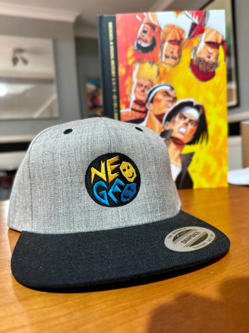 May include: A gray and black baseball cap with a black and yellow embroidered logo that says "NEO GEO" with a smiley face and a video game controller graphic. The cap is a snap back style.