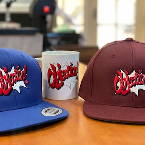 May include: Two blue and maroon baseball caps with a white and red "Objection!" graphic. A white ceramic mug with the same graphic is in the center.