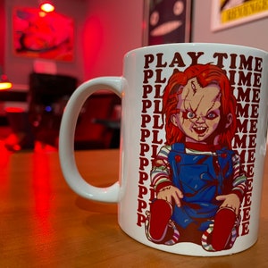 May include: White ceramic mug featuring a red, white and blue illustration of a doll with a menacing expression. The text "PLAY TIME" is repeated multiple times on the mug.