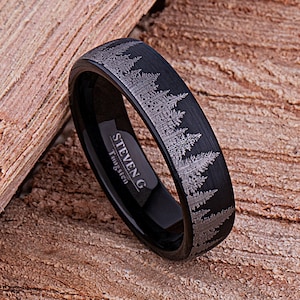 May include: Black tungsten ring with a brushed finish and a silver etched design of a forest tree line. The ring is engraved with "STEVEN G Tungsten".