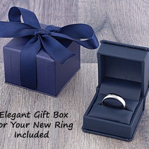 May include: A navy blue ring box with a satin ribbon and bow. The box is open and a silver ring is inside. The text "Elegant Gift Box For Your New Ring Included" is written on a wooden surface.