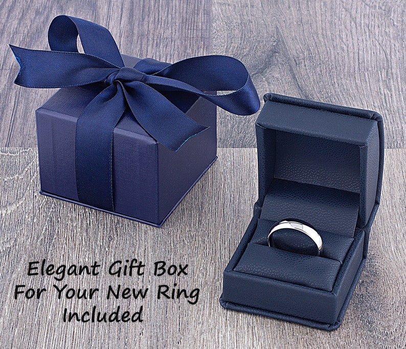 May include: A navy blue ring box with a satin ribbon and bow. The box is open and a silver ring is inside. The text "Elegant Gift Box For Your New Ring Included" is written on a wooden surface.
