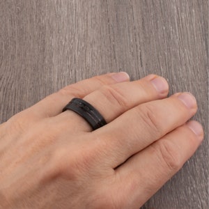 May include: A black and brown ring with a carbon fiber design. The ring is worn on a person's finger.