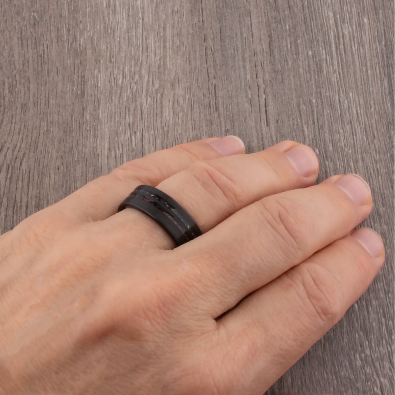 May include: A black and brown ring with a carbon fiber design. The ring is worn on a person's finger.