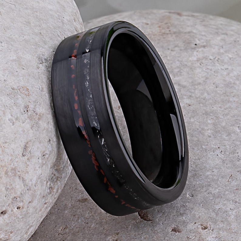 May include: A black tungsten carbide ring with a red and gray inlay. The ring has a polished finish and a beveled edge.