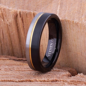 Peut inclure: Une bague en tungstène noire et argent avec une bande dorée. La bague est gravée avec "STEVEN G Tungsten".