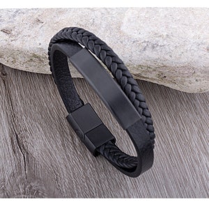 Puede incluir: Pulsera de cuero negro con diseño trenzado y cierre de metal negro. La pulsera tiene una placa de metal rectangular plana.